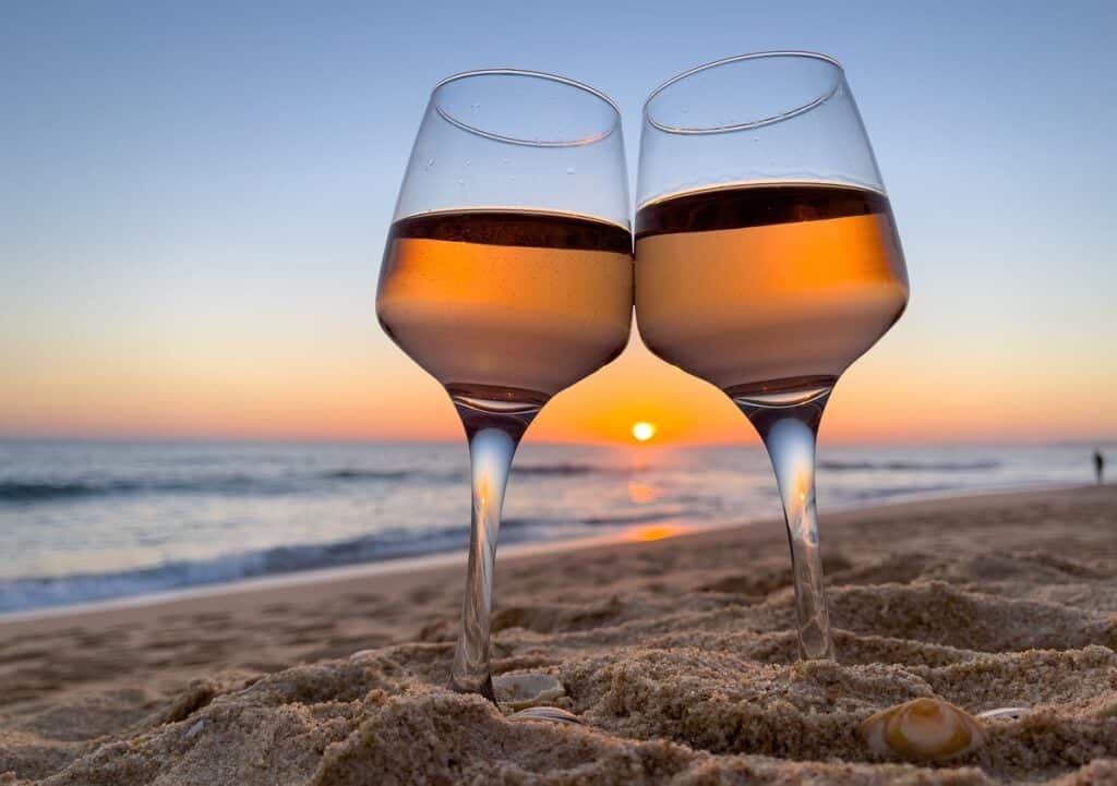 Two glasses of wine resting in the sand by the beach at sunset.